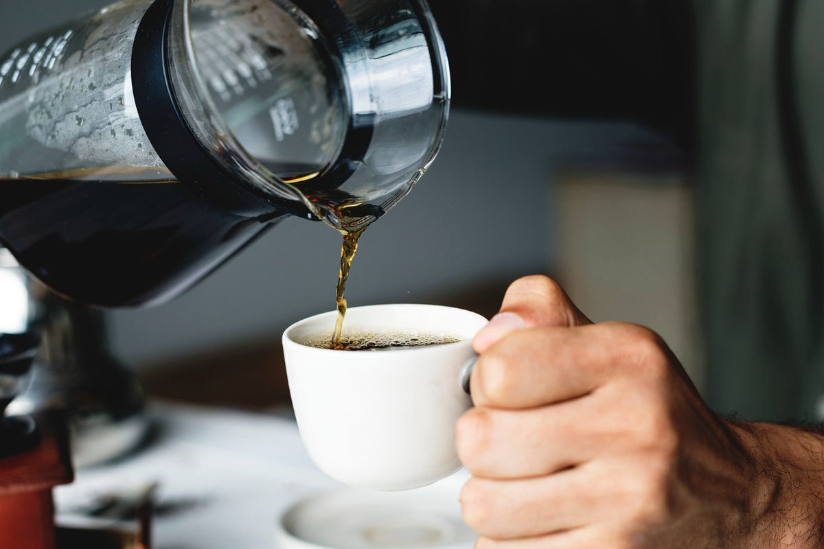 Coffee being poured from a glass and black pour-over style carafe into a white ceramic cup held by a person's hand in a real setting.