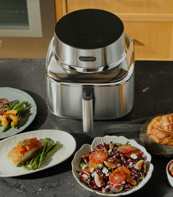 A sleek Cosori air fryer on a kitchen countertop surrounded by plates of freshly cooked food, representing easy everyday cooking.